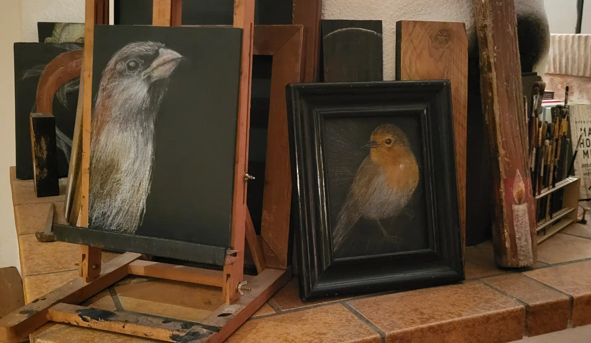 A tabletop in an artist’s workspace displays several small paintings and wooden panels. Two bird paintings are most prominent—one on a small wooden easel showing a brown-and-white bird with a pinkish head, and another in a black frame showing a bird with orange and brown plumage. Additional unfinished boards, brushes, and art supplies surround them, creating a casual studio atmosphere.